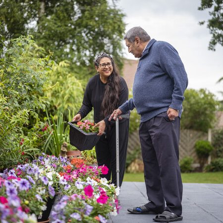 A woman helping a man with a walking stick in the garden.